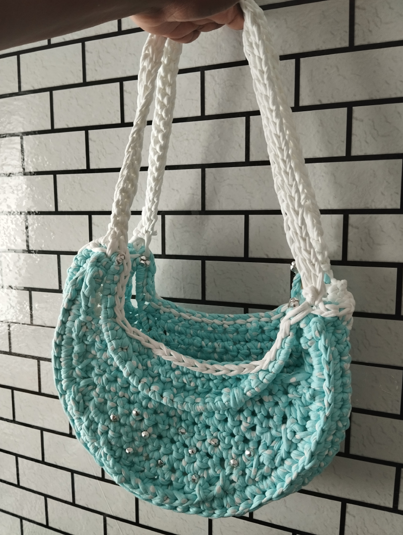 Un sac à main bleu clair crocheté à la main, orné de petites perles argentées et doté de anses blanches, est accroché contre un mur de briques noires et blanches.