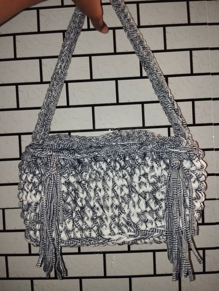 Une main tient un sac à main tressé bleu et blanc contre un mur carrelé. Le sac est doté d'anses tressées et de longs pompons, lui conférant un style à la fois décontracté et raffiné.